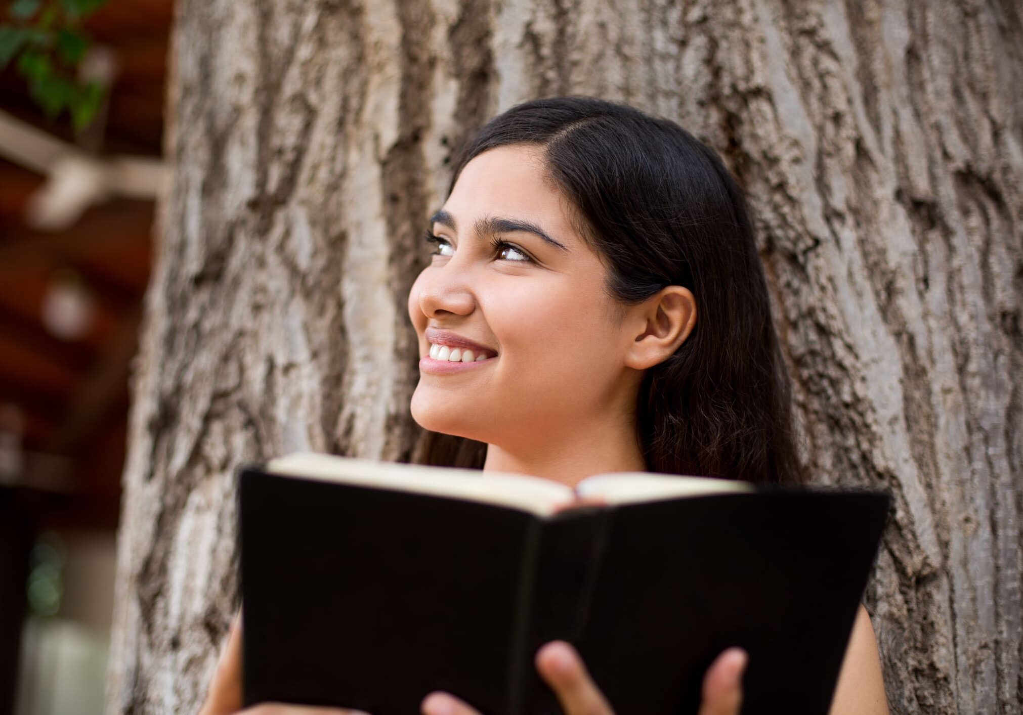 Woman smiling while reading book under tree.